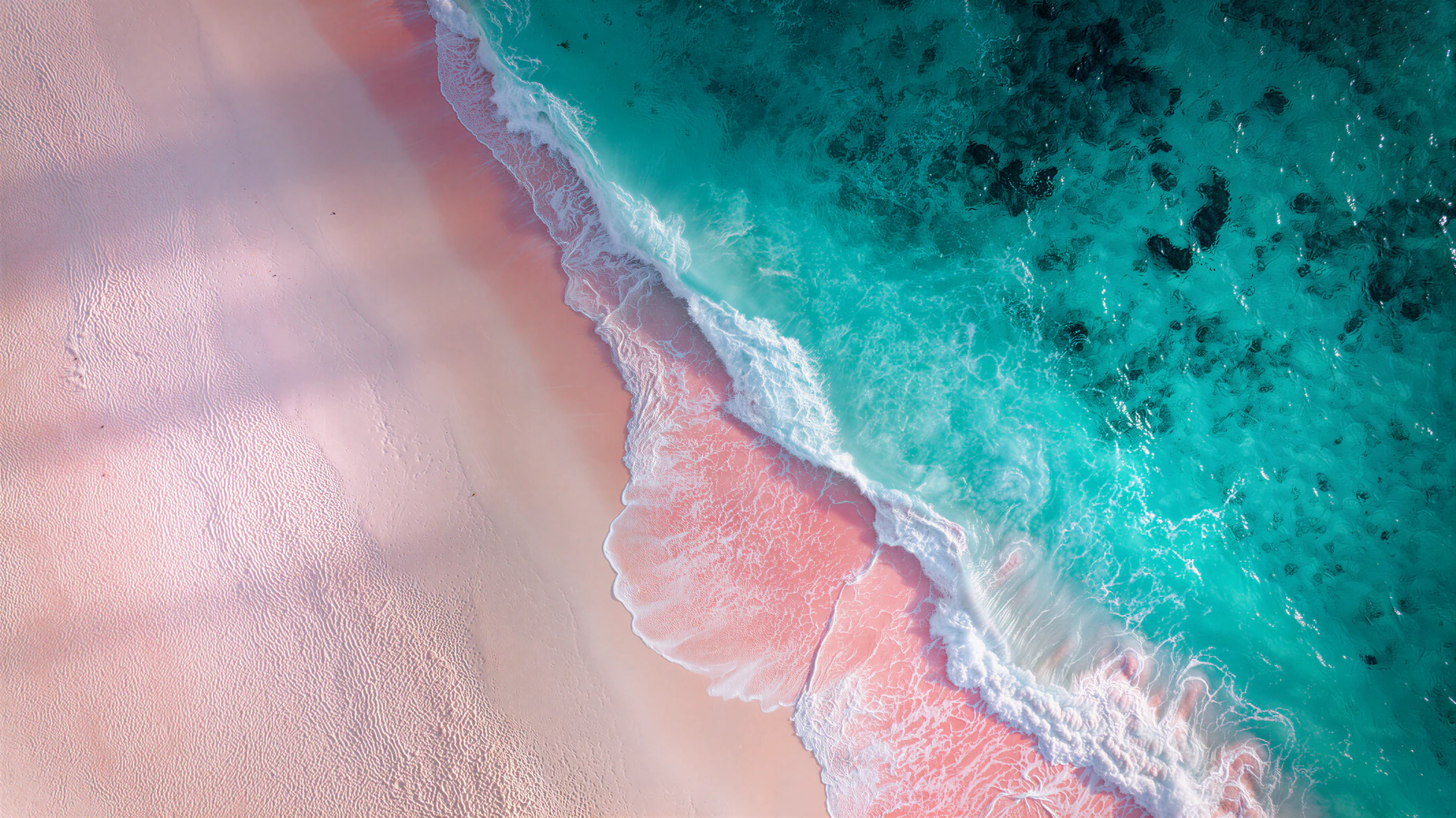 An aerial view of turquoise seawater sweeping across a dreamy pink sand beach. Foamy waves create a vibrant, abstract coastal landscape.