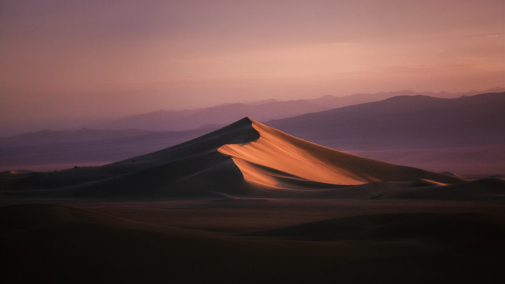A lone crescent dune catches the last ember of twilight above a vast, shadowed desert. Muted mountains and rose-tinted skies create a cinematic, tranquil scene.