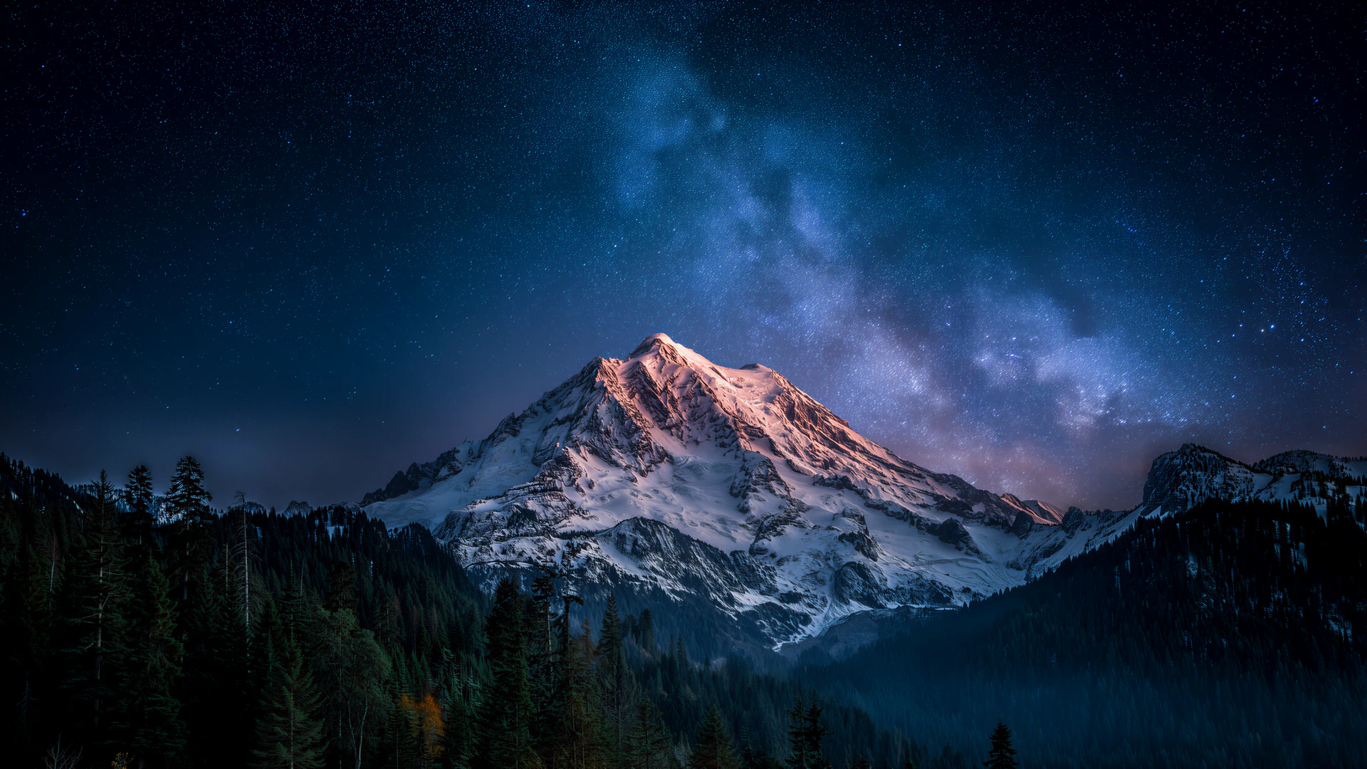 A snow-capped mountain glows beneath the Milky Way, rising above a shadowy evergreen forest. Cool midnight blues meet warm alpine light in this cinematic nig…